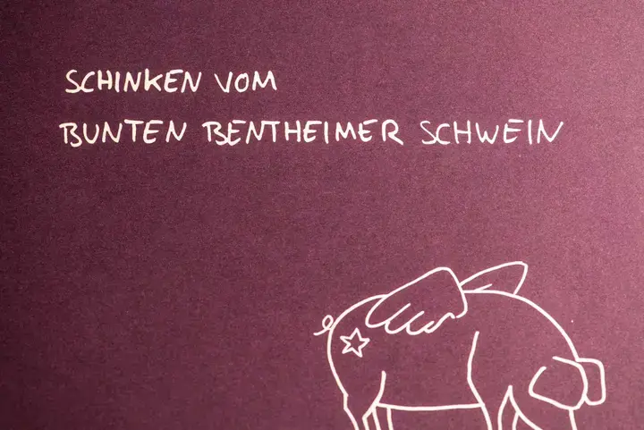 Schriftzug "Schinken vom bunten Bentheimer Schwein" über beflügelter Sau in der Optik eines Reisepasses